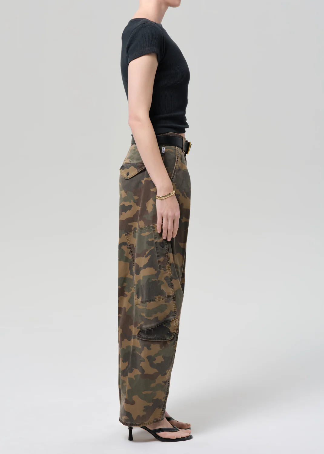 Person stands in profile against a plain background, wearing a fitted black short-sleeve top, Citizens Of Humanity/AGOLDE flight pant in camo/Khaki, a black belt, gold bracelet, and black heeled sandals.
