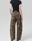 A person faces away from the camera wearing a black short-sleeve top, Citizens Of Humanity/AGOLDE flight pant in camo/Khaki, and heeled sandals against a plain light background.