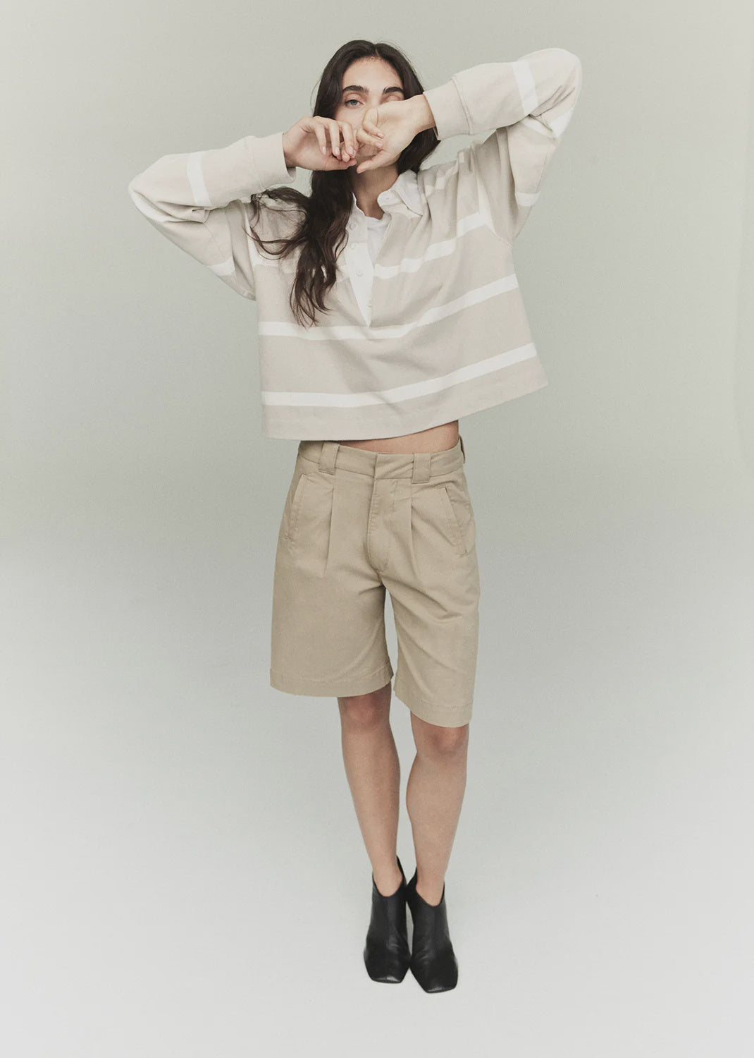 A person wearing the Cara Pleated Short by Citizens Of Humanity/AGOLDE, a beige and white striped sweater, and black ankle boots stands against a plain background. With long hair, they playfully cover their mouth with the sweater's sleeve, giving off a vintage vibe.