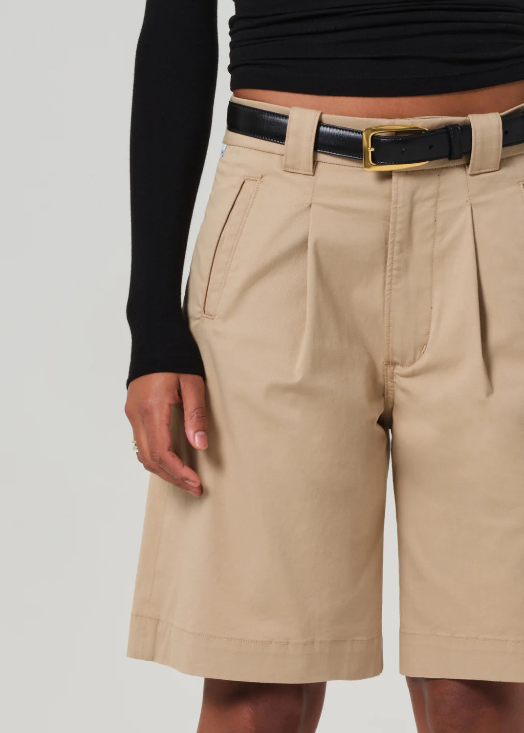 Dressed in a black long-sleeve top and Cara Pleated Shorts by Citizens Of Humanity/AGOLDE in beige lightweight twill, with a gold-buckled black belt enhancing the waist, this look seamlessly blends textures on the upper legs and visible arm.