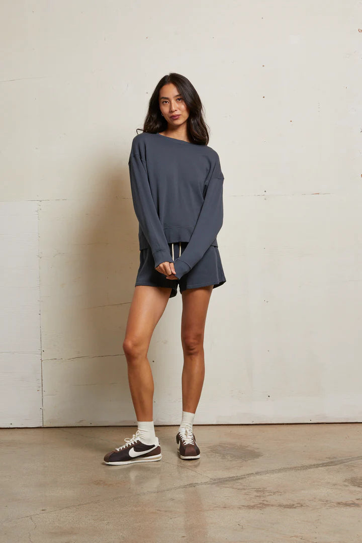 A woman with long dark hair poses casually indoors against a plain wall, wearing the Perfectwhitetee French Terry Sweatshirt and matching shorts in dark gray, paired with white socks and brown Nike sneakers.