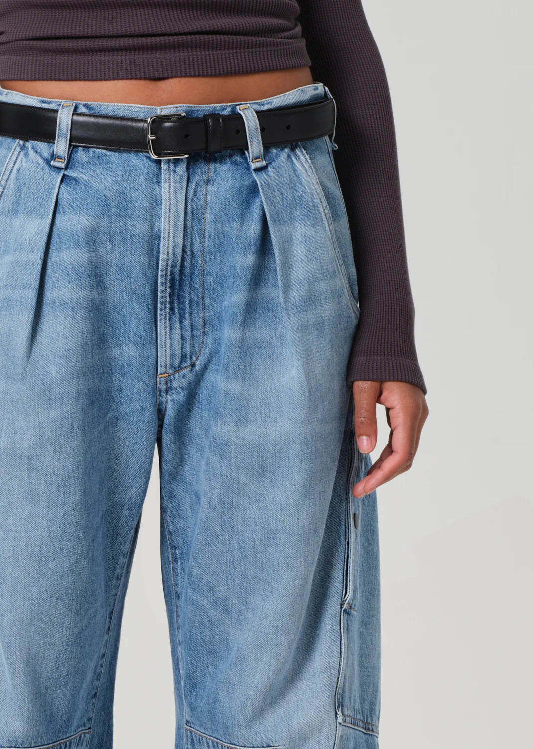 The image shows someone in Citizens Of Humanity/AGOLDE's Flight Pant, high-waisted blue jeans with pleats, paired with a long-sleeve dark ribbed top and a black belt. With one arm hanging casually by their side, the vintage-inspired utility look blends style and sustainability.