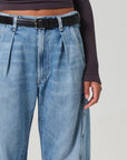 The image shows someone in Citizens Of Humanity/AGOLDE's Flight Pant, high-waisted blue jeans with pleats, paired with a long-sleeve dark ribbed top and a black belt. With one arm hanging casually by their side, the vintage-inspired utility look blends style and sustainability.