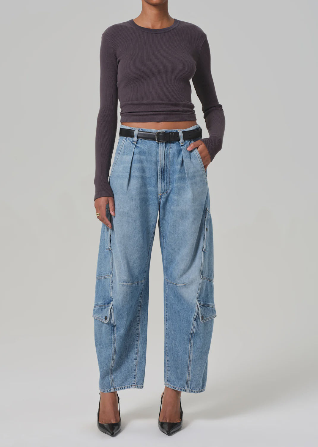 Against a plain backdrop, a model showcases their vintage-inspired utility style wearing AGOLDE's Flight Pant. The look features light blue cargo jeans made from regenerative cotton, paired with a fitted black top, black pointed heels, and a matching belt.