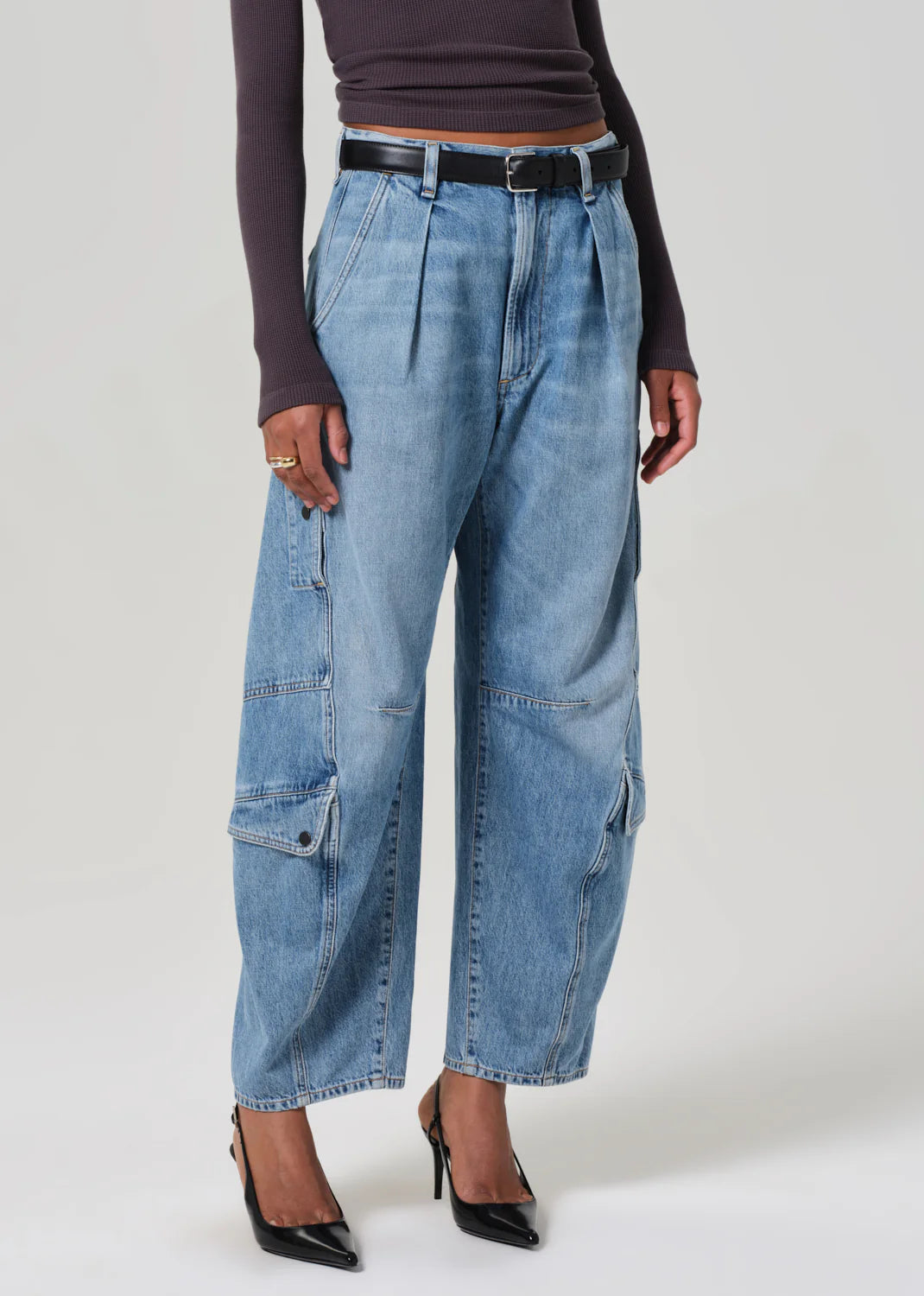 Someone wears Citizens Of Humanity/AGOLDE's Flight Pant, featuring vintage-inspired utility design with high-waisted, wide-legged blue denim cargo styling and a black belt. They're paired with a dark long-sleeve top and black pointed heels against a light gray background.