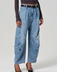 Someone wears Citizens Of Humanity/AGOLDE's Flight Pant, featuring vintage-inspired utility design with high-waisted, wide-legged blue denim cargo styling and a black belt. They're paired with a dark long-sleeve top and black pointed heels against a light gray background.