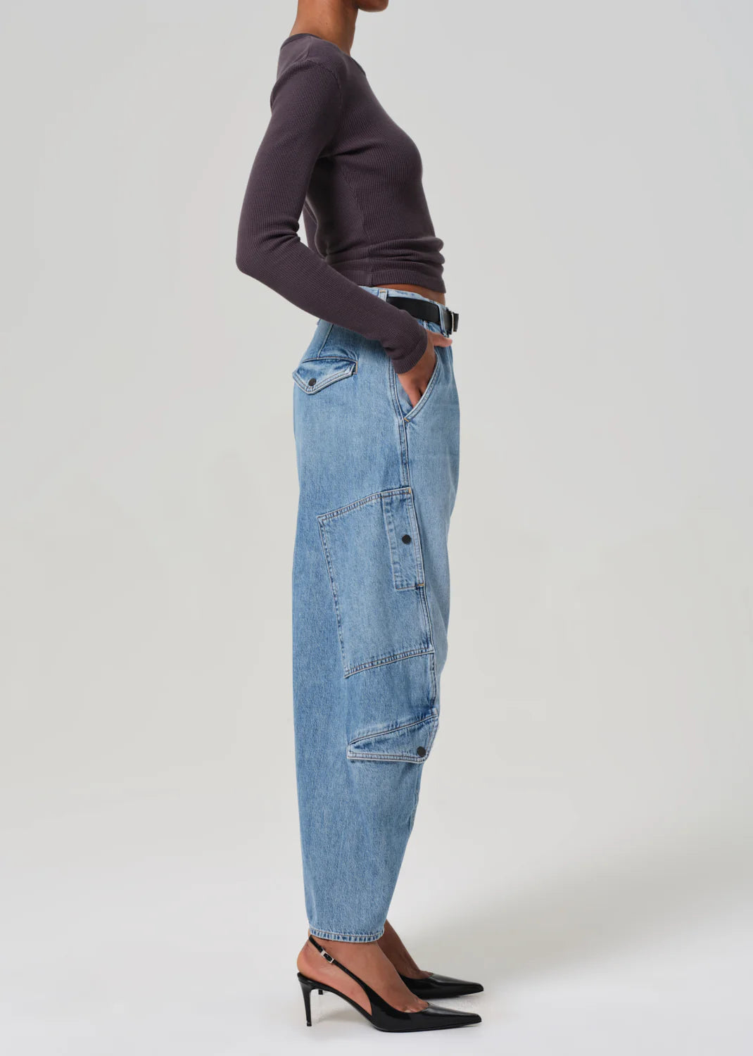 A person in profile against a plain background wears Citizens Of Humanity/AGOLDE Flight Pant—a long-sleeve black top and loose-fitting blue jeans with a vintage utility fit. The jeans, made of regenerative cotton, feature a large side pocket. Black pointed heels complete the look, with hands in pockets.