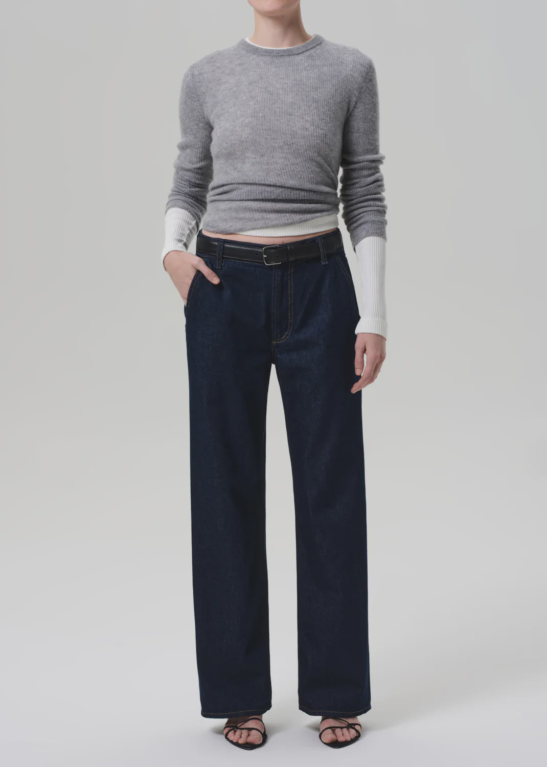 A person stands against a plain light background wearing Citizens Of Humanity Annina Pocket Trouser wide-leg jeans in dark indigo denim, paired with a fitted grey sweater with white sleeves and black sandals. Their face is not visible.
