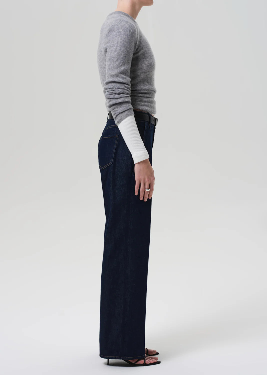 A person stands in profile against a plain background, wearing a fitted grey long-sleeve sweater, Citizens Of Humanity Annina Pocket Trouser in dark indigo denim, and black open-toe sandals.