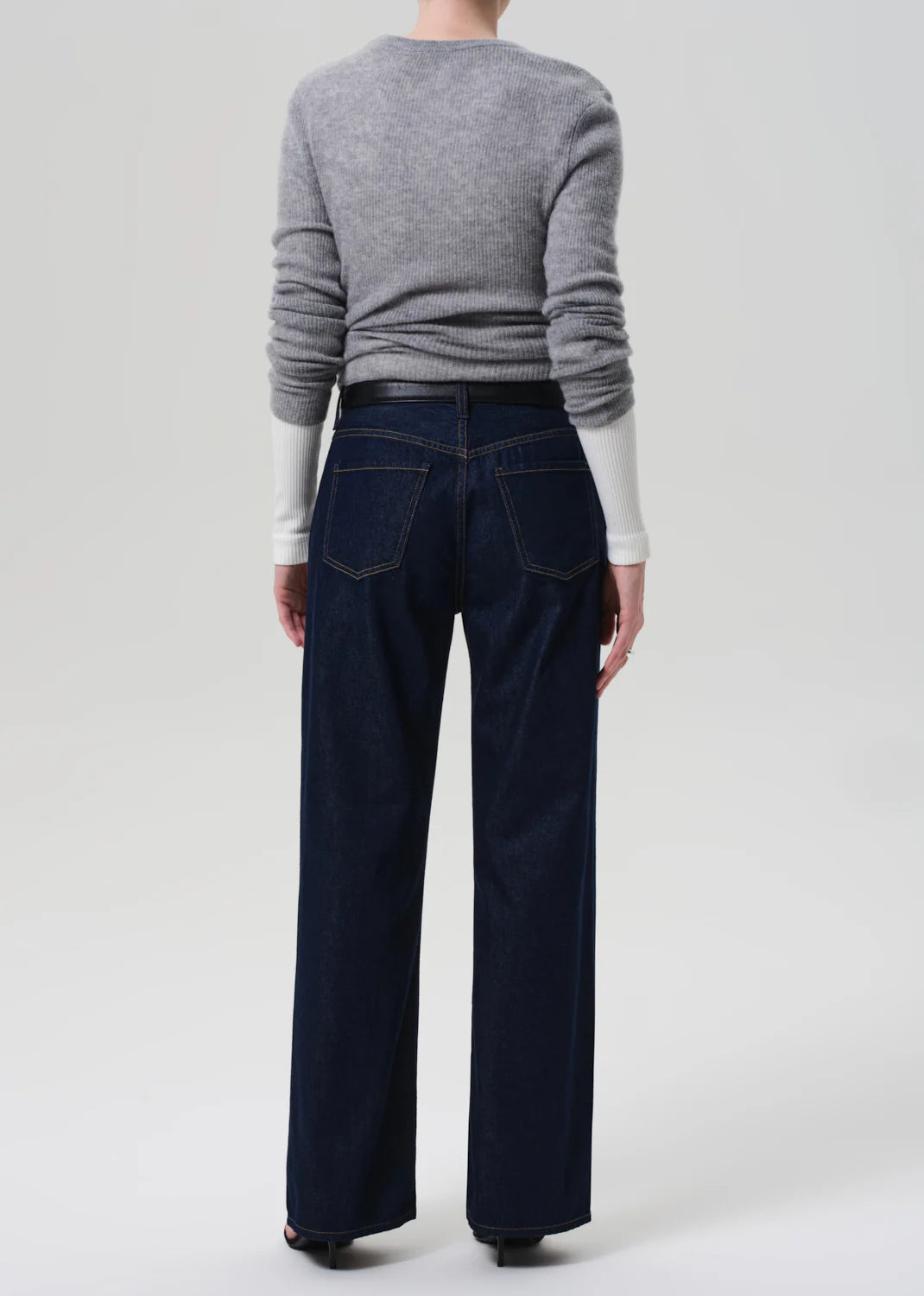 A person faces away from the camera, wearing a gray ribbed sweater over a white long-sleeve top and Citizens Of Humanity Annina Pocket Trouser in dark indigo—a high-rise, wide-leg denim style—against a plain light background.