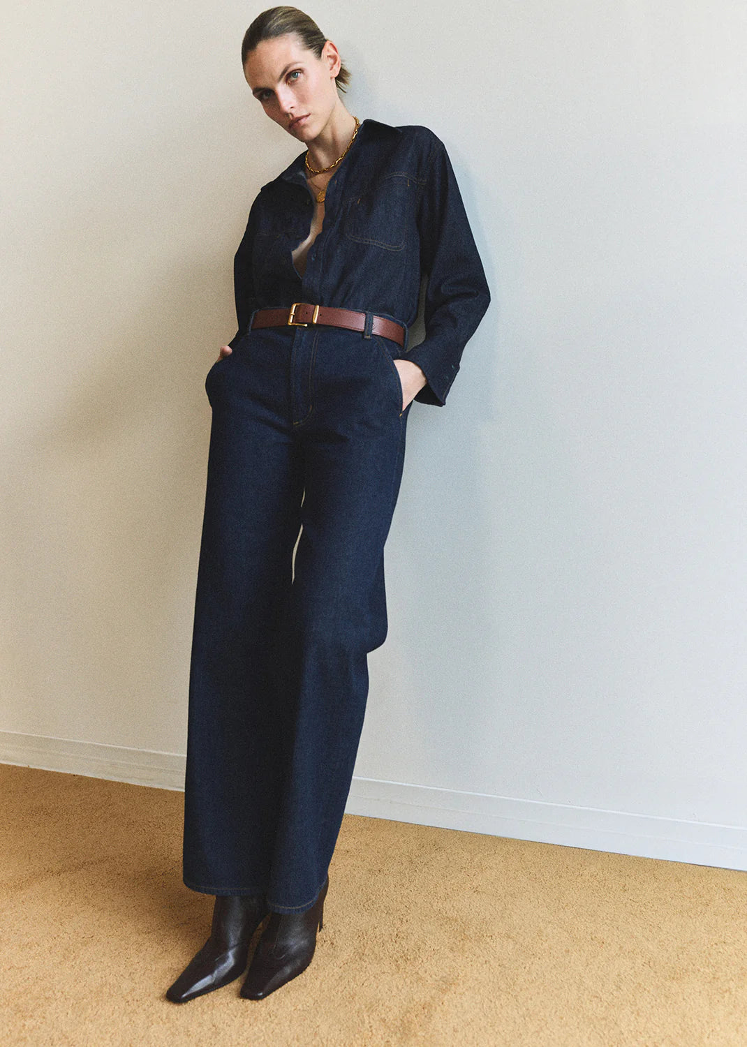 A person stands against a white wall wearing the Annina Pocket Trouser by Citizens Of Humanity/AGOLDE, crafted from recycled cotton. Styled with a brown belt and pointed-toe boots, they pose casually with hands in pockets.