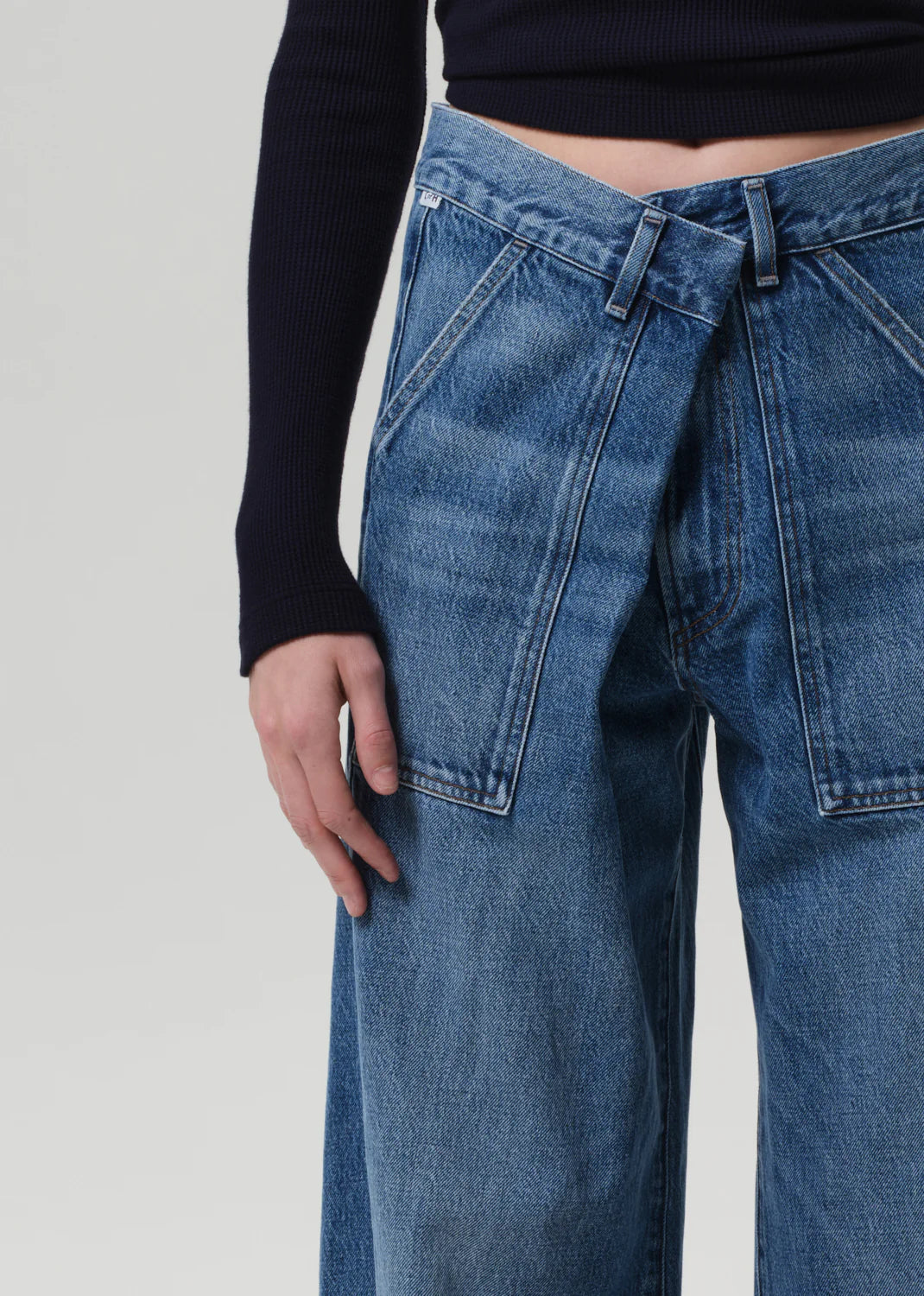 A person wearing Citizens Of Humanity/AGOLDE's Ines Utility Trouser in medium indigo denim with an overlapping waistband and a black ribbed long-sleeve top, shown from mid-torso to mid-thigh against a plain background.