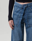 A person wearing Citizens Of Humanity/AGOLDE's Ines Utility Trouser in medium indigo denim with an overlapping waistband and a black ribbed long-sleeve top, shown from mid-torso to mid-thigh against a plain background.