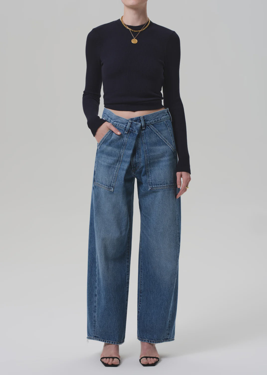 A person wears a black long-sleeve crop top, Citizens Of Humanity/AGOLDE Ines Utility Trouser in medium indigo regenerative cotton denim with front pockets and tied belt, black heeled sandals, and gold jewelry against a plain light background.