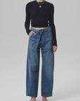 A person wears a black long-sleeve crop top, Citizens Of Humanity/AGOLDE Ines Utility Trouser in medium indigo regenerative cotton denim with front pockets and tied belt, black heeled sandals, and gold jewelry against a plain light background.