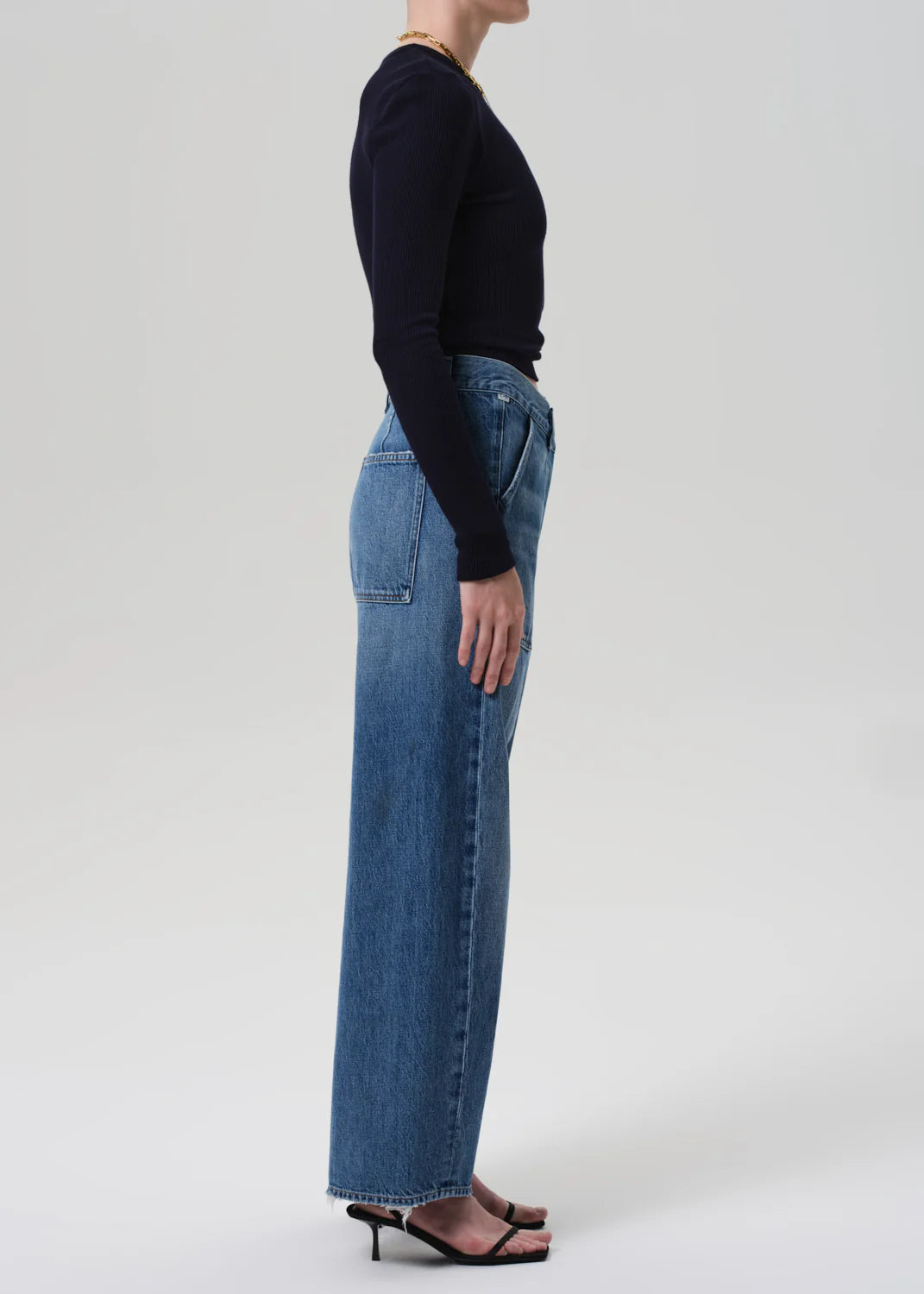 A person stands in profile against a plain background, wearing a long-sleeve black top, Citizens Of Humanity/AGOLDE Ines Utility Trouser in medium indigo denim, and black heeled sandals.