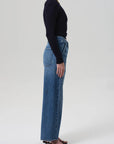 A person stands in profile against a plain background, wearing a long-sleeve black top, Citizens Of Humanity/AGOLDE Ines Utility Trouser in medium indigo denim, and black heeled sandals.