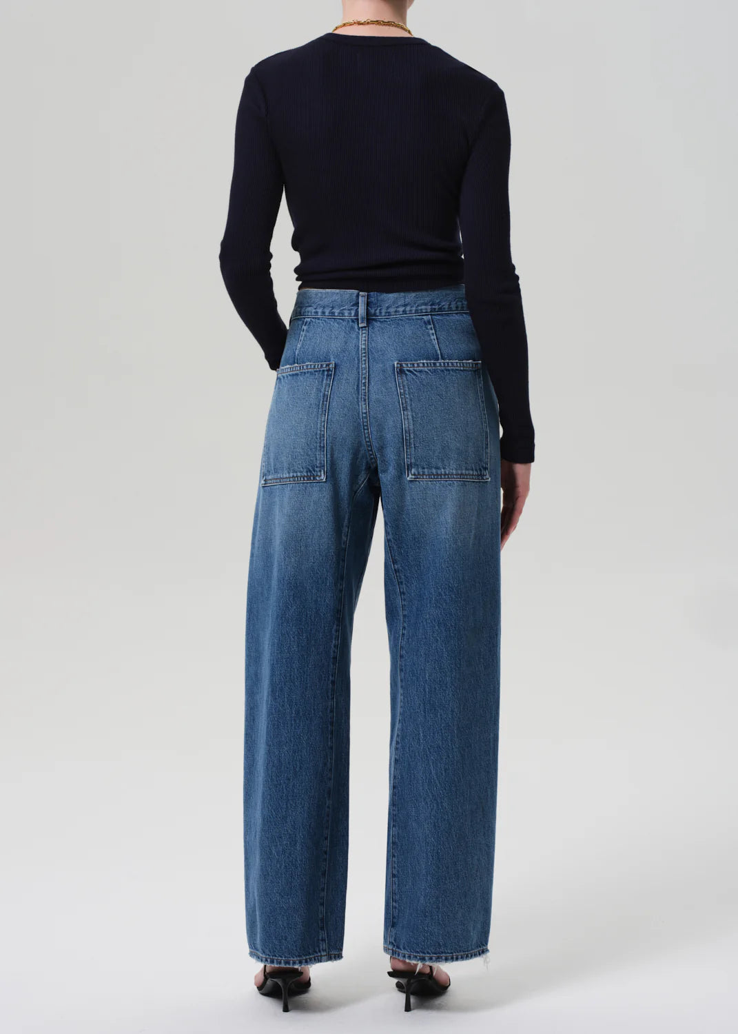 A person stands facing away from the camera, wearing a black long-sleeve top and Citizens Of Humanity/AGOLDE Ines Utility Trouser jeans in medium indigo denim with large back pockets, paired with black heeled sandals against a plain light background.