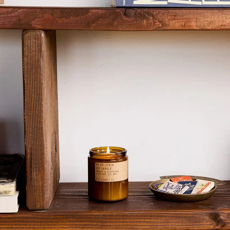 A lit PFC Candle by Faire in a brown glass container sits on a wooden shelf next to a dish of matchbooks, with books stacked below and at the upper right edge of the image.