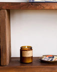 A lit PFC Candle by Faire in a brown glass container sits on a wooden shelf next to a dish of matchbooks, with books stacked below and at the upper right edge of the image.