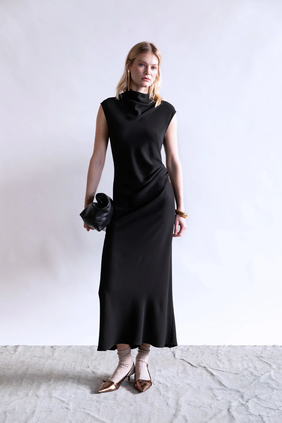 A woman stands against a plain light background in the Psophia Naia Drape Dress—sleeveless, long, and black—paired with brown flats, ankle socks, a black clutch bag, and gold earrings and bracelets.