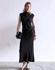 A woman stands against a plain light background in the Psophia Naia Drape Dress—sleeveless, long, and black—paired with brown flats, ankle socks, a black clutch bag, and gold earrings and bracelets.