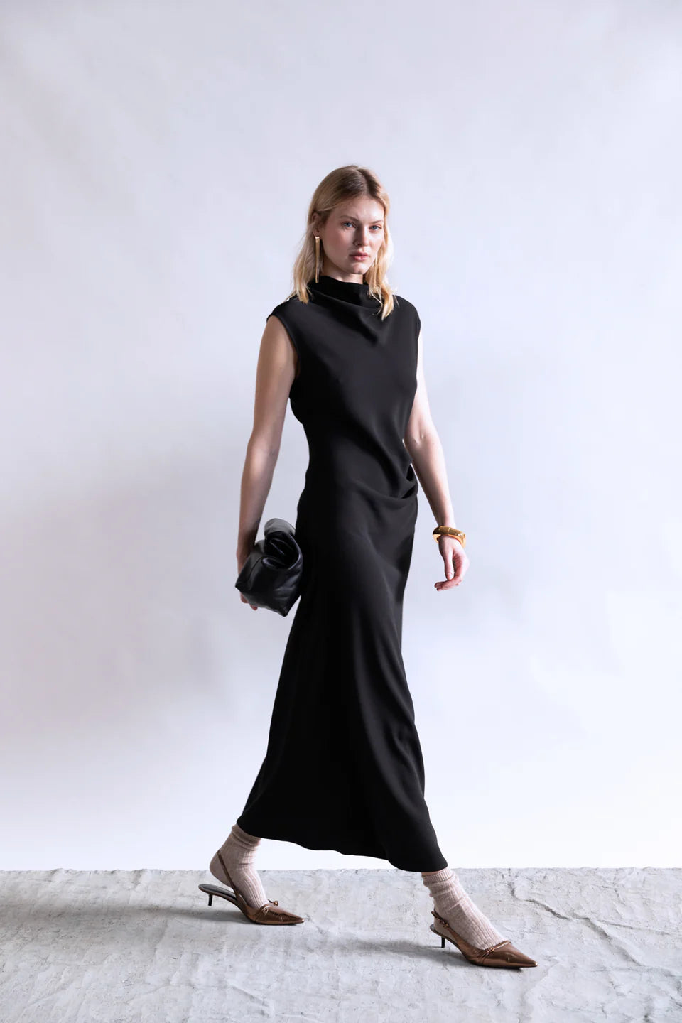 A woman wears the Psophia Naia Drape Dress, a sleeveless, ankle-length minimal piece, against a plain white background. She carries a black clutch, pairs heeled shoes with light socks, and accessorizes with gold bracelets.