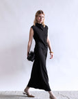 A woman wears the Psophia Naia Drape Dress, a sleeveless, ankle-length minimal piece, against a plain white background. She carries a black clutch, pairs heeled shoes with light socks, and accessorizes with gold bracelets.