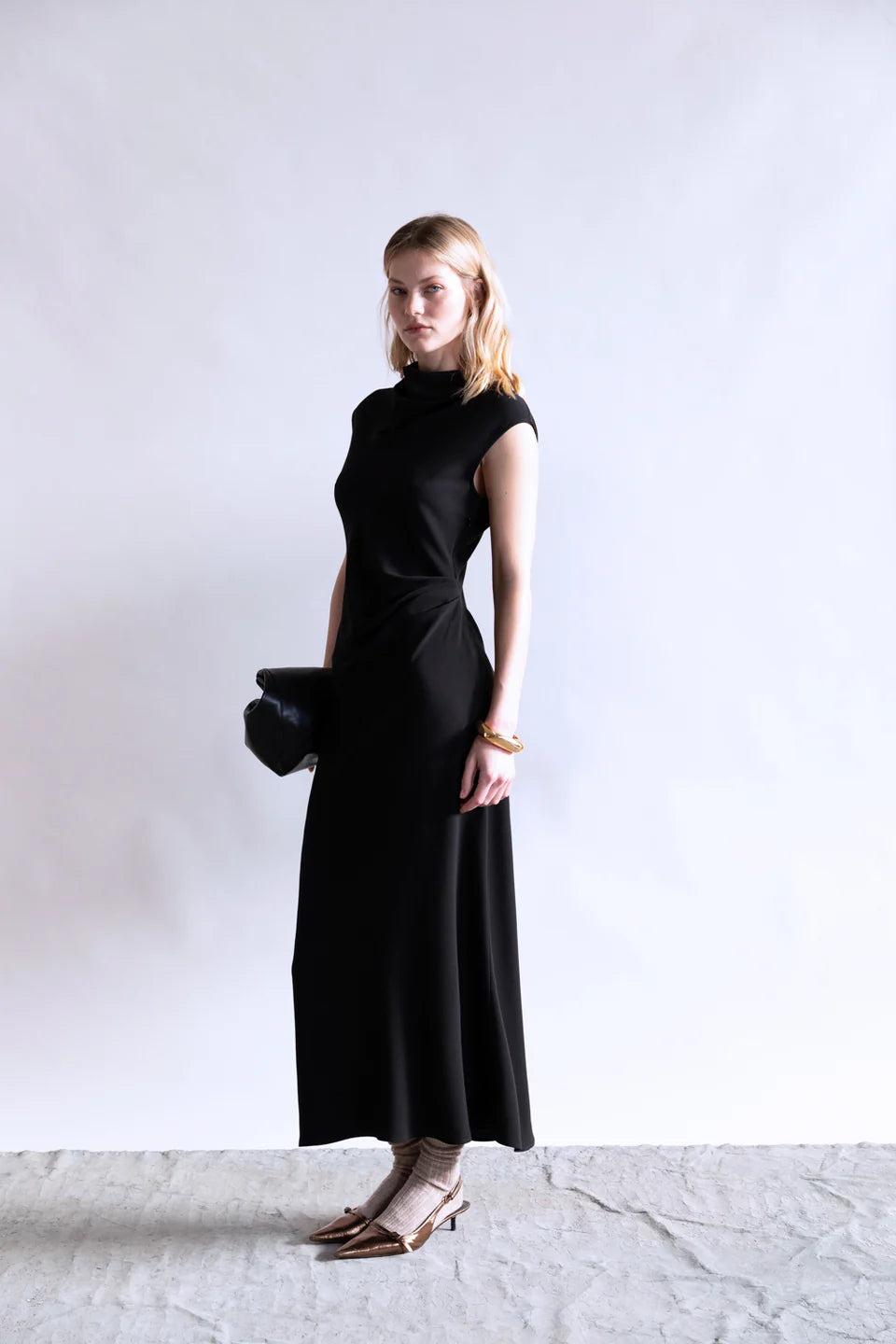 A woman with shoulder-length blonde hair stands against a plain background, wearing the Psophia Naia Drape Dress in black, gold shoes with socks, and holding a black clutch. Her expression is neutral.