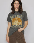 A person with long dark hair is wearing a People Of Leisure Lainey Wilson Heart Like A Truck shirt, crafted from organic cotton, along with brown pants. The dark gray tee displays the phrase "I Got A Heart Like a Truck" above an illustration of a pickup truck. They are standing against a plain background.