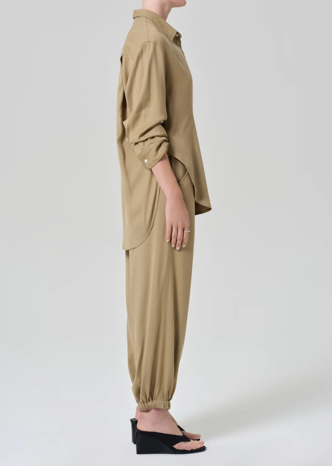 A person stands in profile wearing Citizens Of Humanity/AGOLDE's bubble pull on in portofino twill—light brown, loose-fit drawstring pants—paired with a light shirt and black open-toe wedge sandals against a plain light background.