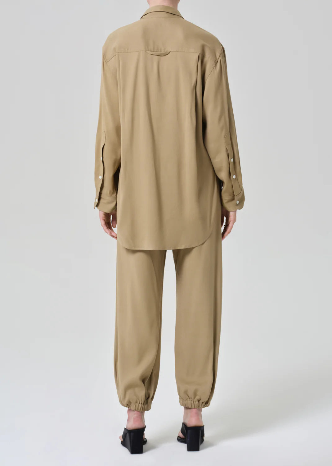 A person faces away, wearing the Citizens Of Humanity/AGOLDE bubble pull on in portofino twill—loose-fitting pants with elastic cuffs and a drawstring waist—styled with a beige long-sleeve shirt and black heeled sandals against a plain light background.