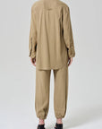 A person faces away, wearing the Citizens Of Humanity/AGOLDE bubble pull on in portofino twill—loose-fitting pants with elastic cuffs and a drawstring waist—styled with a beige long-sleeve shirt and black heeled sandals against a plain light background.