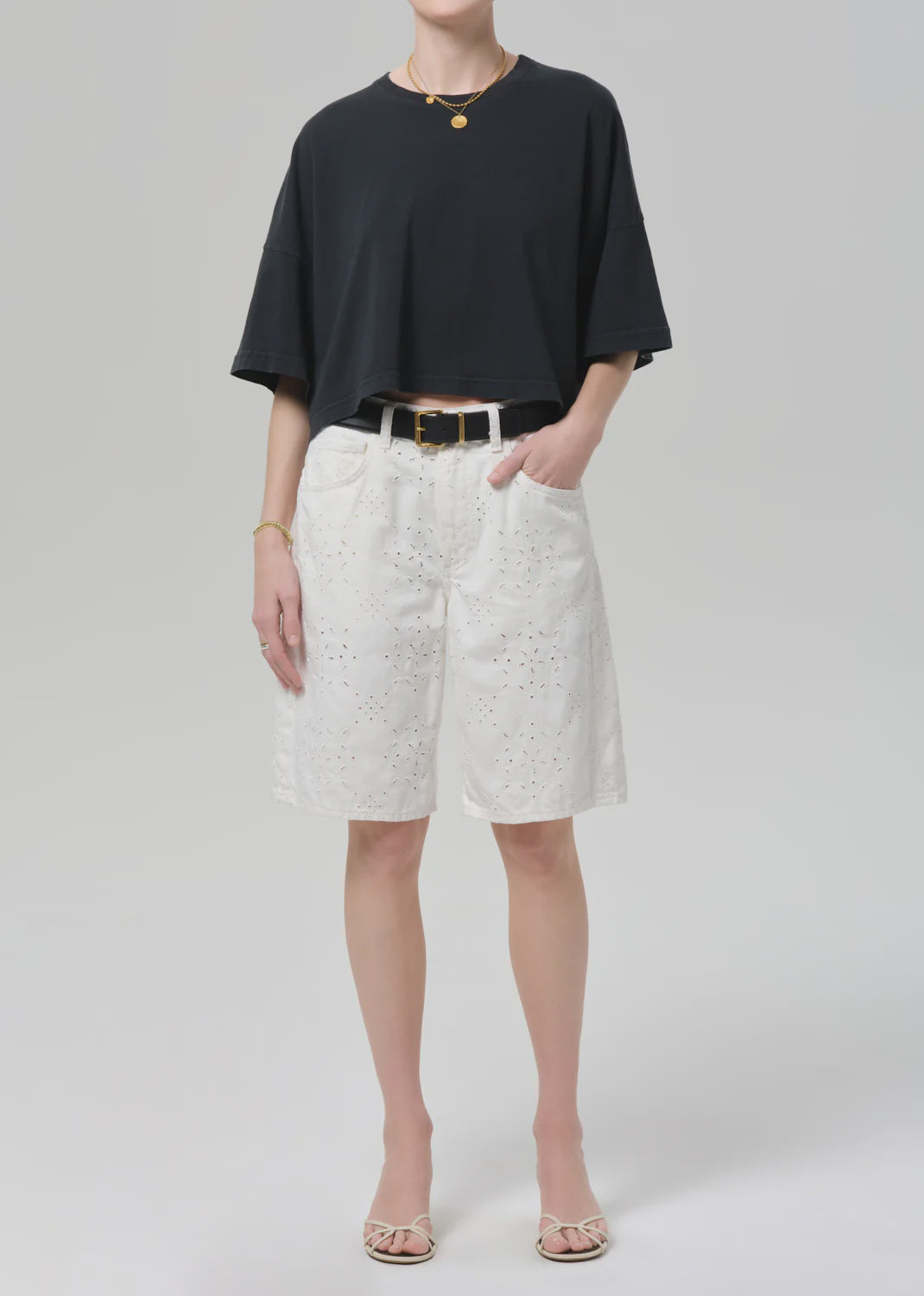 A person wears the Citizens Of Humanity/AGOLDE court denim short in blanc with a loose black T-shirt, a black belt, open-toe sandals, and simple jewelry, standing against a plain light gray background.