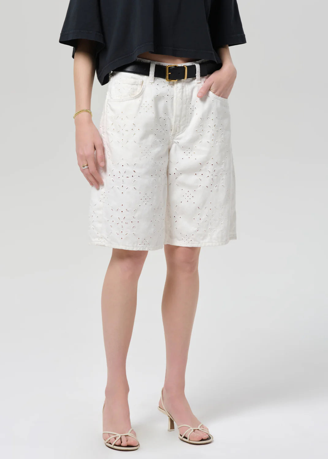 A person wears a black top, Citizens Of Humanity/AGOLDE court denim short in blanc with a black belt, and strappy heeled sandals, standing with one hand in a pocket against a plain background.