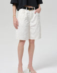 A person wears a black top, Citizens Of Humanity/AGOLDE court denim short in blanc with a black belt, and strappy heeled sandals, standing with one hand in a pocket against a plain background.
