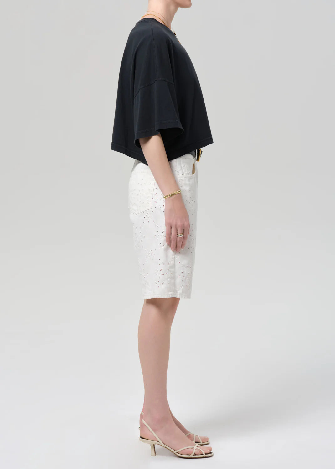 A woman stands in profile against a plain background, wearing a loose black short-sleeve top, Citizens Of Humanity/AGOLDE court denim short in blanc, strappy nude heels, and minimal jewelry.