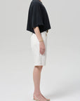 A woman stands in profile against a plain background, wearing a loose black short-sleeve top, Citizens Of Humanity/AGOLDE court denim short in blanc, strappy nude heels, and minimal jewelry.