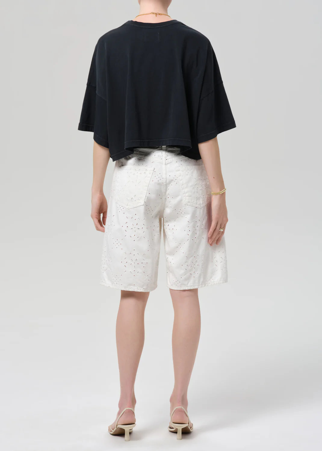 A person stands facing away, wearing a loose black short-sleeve top, Citizens Of Humanity/AGOLDE court denim short in blanc, and light-colored heeled sandals against a plain background.