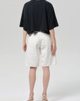 A person stands facing away, wearing a loose black short-sleeve top, Citizens Of Humanity/AGOLDE court denim short in blanc, and light-colored heeled sandals against a plain background.