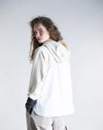 A person with long curly hair wears the Psophia Cropped Hoodie W/ Shirt Hem, featuring beige sleeves, paired with light gray pants. They stand against a plain light background, facing away and looking back over their shoulder.