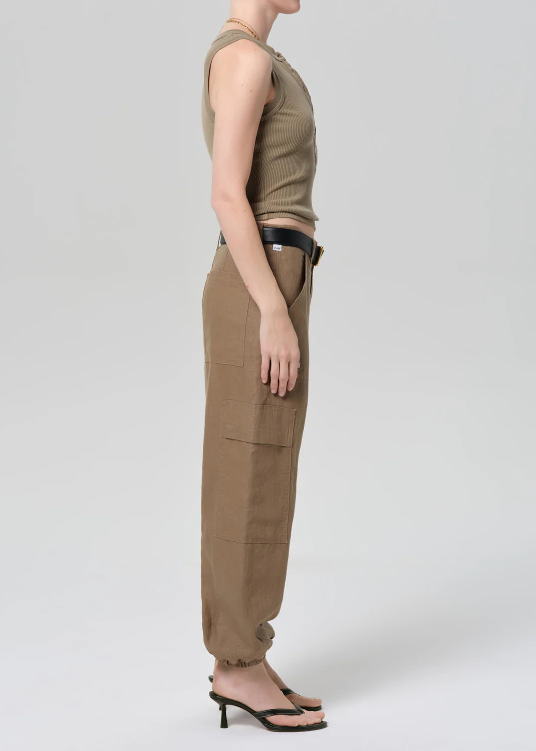 A person stands in profile against a plain background, wearing the ciela balloon utility pants by Citizens Of Humanity/AGOLDE with a sleeveless taupe top, black belt, and heeled sandals. Their hair is pulled back and arms rest at their sides.
