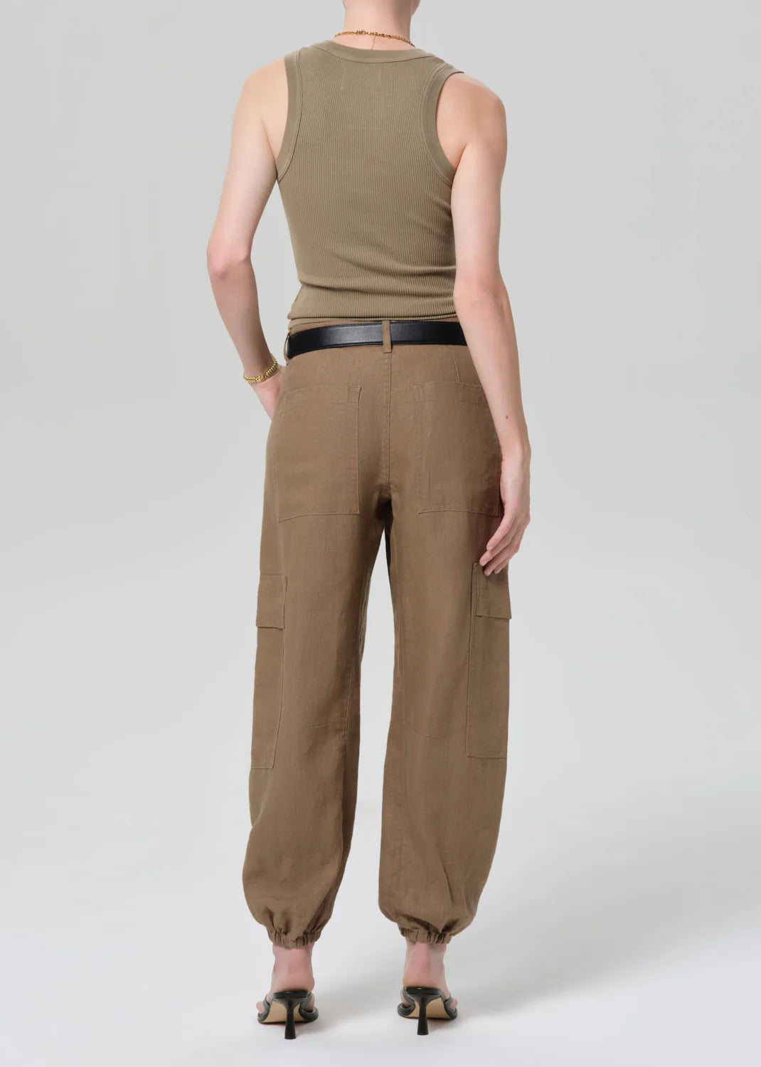 A person faces away, wearing Citizens Of Humanity/AGOLDE ciela balloon utility pants with cargo pockets and cuffed ankles, a fitted khaki tank top, black belt, heeled sandals, and a bracelet. The background is plain light gray.
