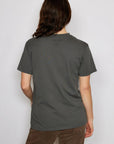 A person with long brown hair wearing a dark gray unisex t-shirt by People Of Leisure and brown pants is seen from behind against a plain gray background.