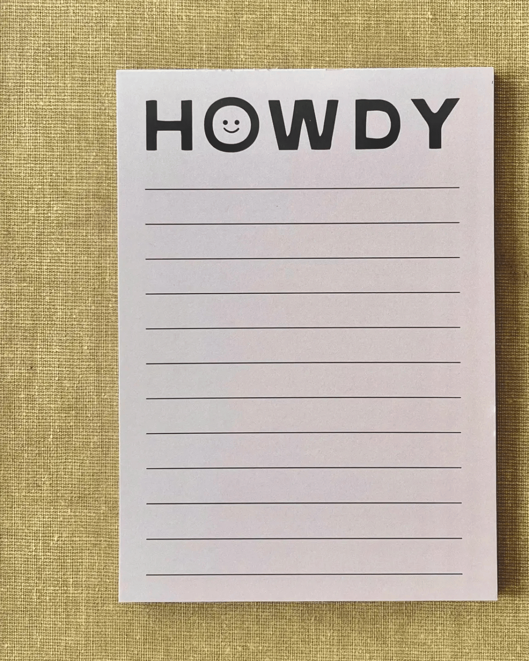 The Faire Howdy Note Pad, adorned with a "HOWDY" header and a smiling face, brings a charming touch to jotting down notes or pecan pie recipes. It includes ruled lines for easy writing against a textured, neutral-toned fabric background.