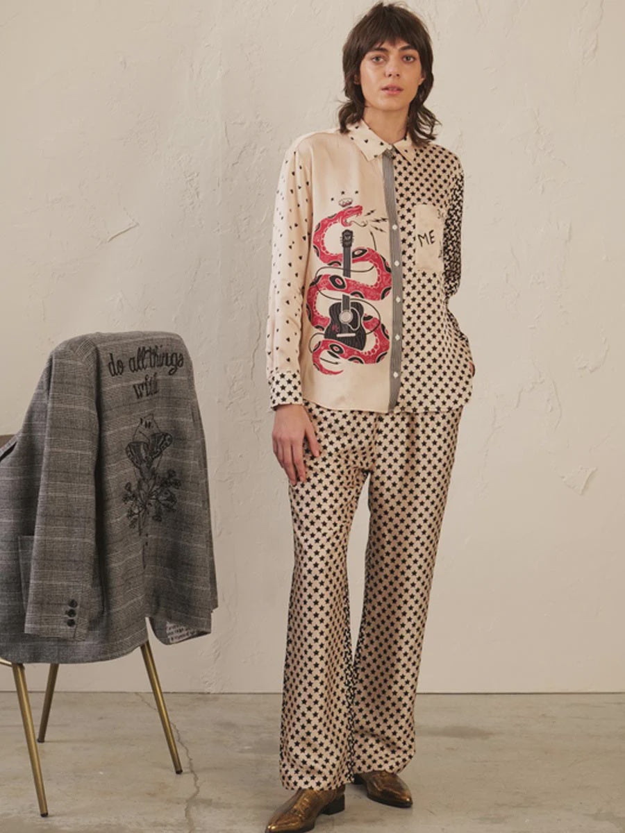 A person stands by a plain wall wearing Me369's Marilyn Trousers with a matching shirt featuring a red snake and guitar graphic. A gray jacket printed with "do all things with" is draped over a nearby chair.