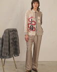 A person stands by a plain wall wearing Me369's Marilyn Trousers with a matching shirt featuring a red snake and guitar graphic. A gray jacket printed with "do all things with" is draped over a nearby chair.