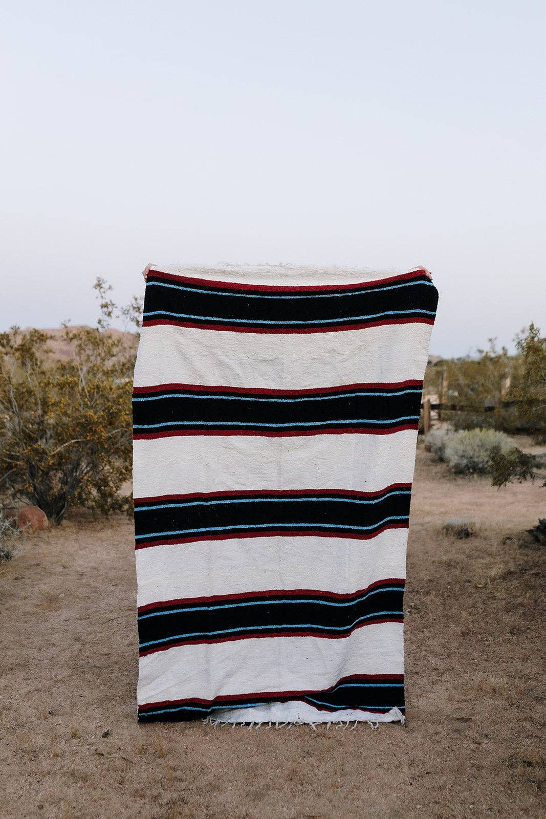 Handwoven Canyon Creek blanket by Bungalow Furniture standing upright on sand with bushes in the background.