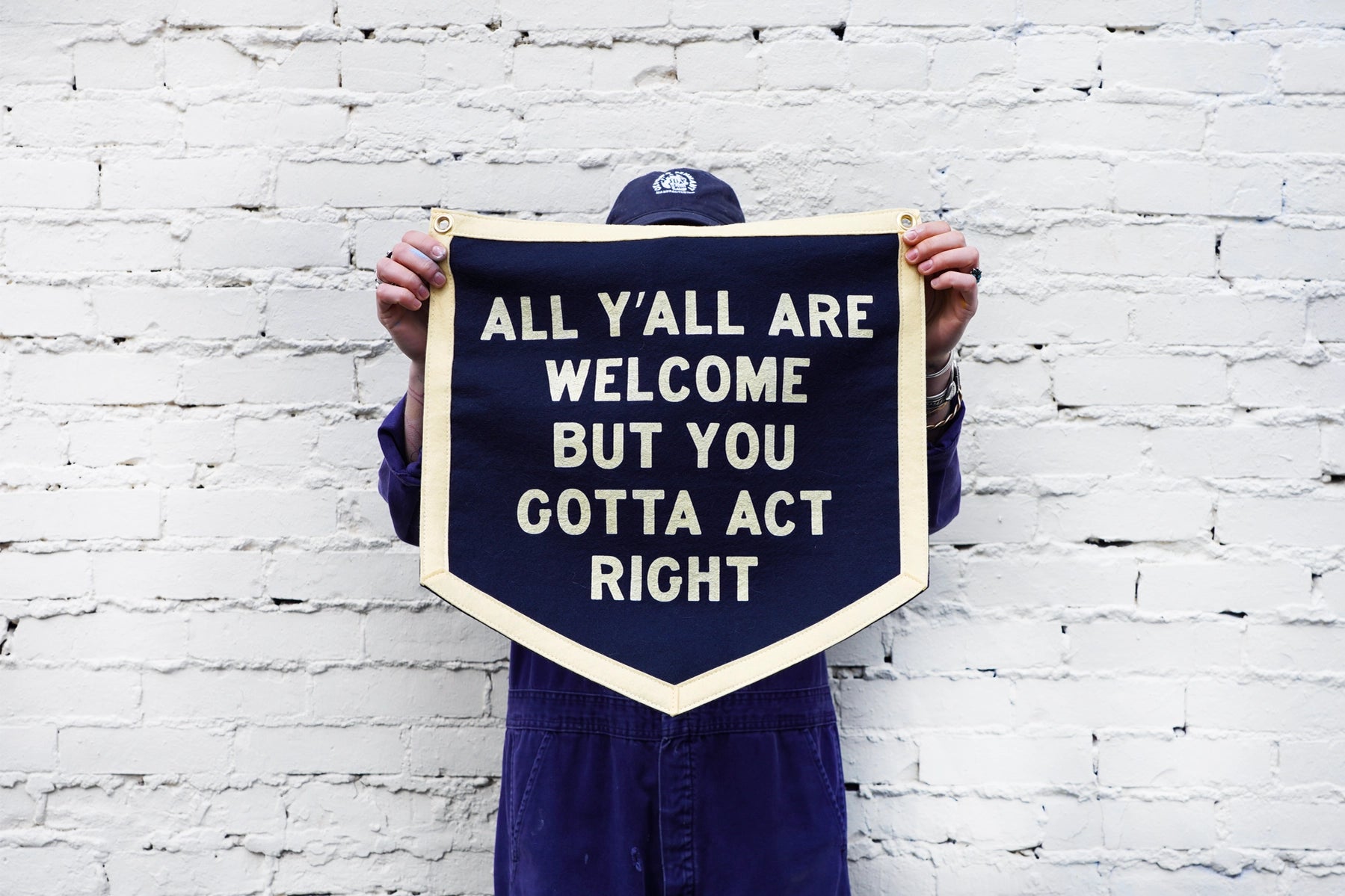 In Rockwall, TX, a person in a purple outfit and cap stands against a white brick wall holding Oxford Pennant's "All Y'All Are Welcome..." navy banner.