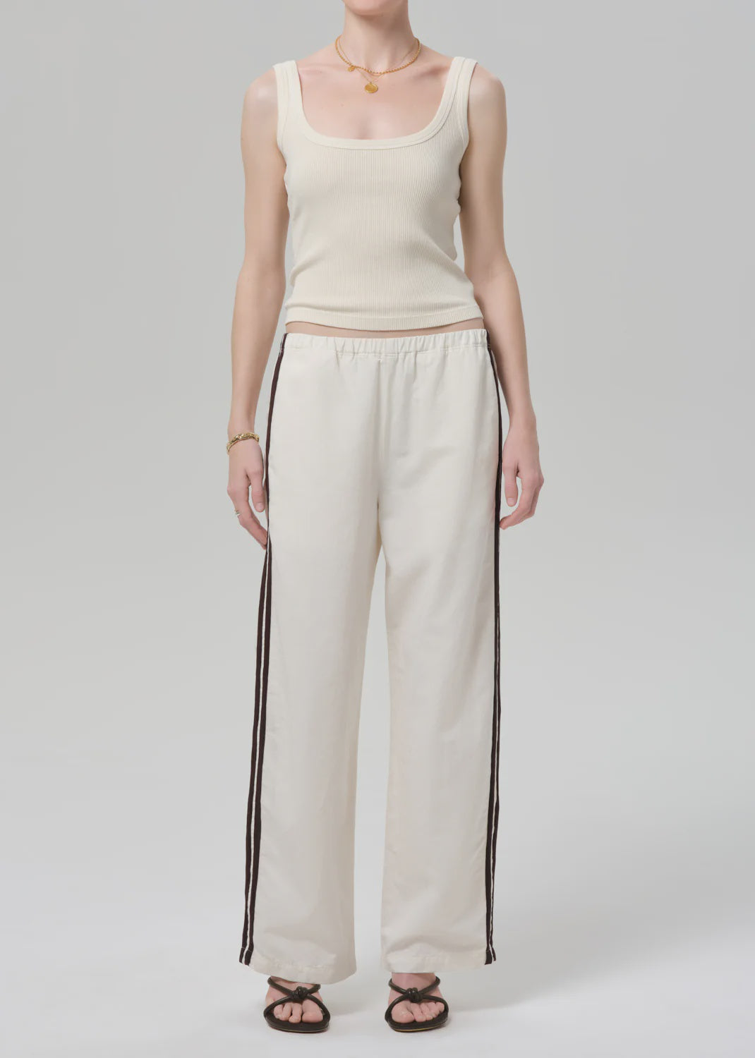 A person stands against a plain background wearing an off white Citizens Of Humanity/AGOLDE aurora track pant with a brown stripe, a cream ribbed tank top, black sandals, a gold necklace, and a bracelet. Their face is not shown.